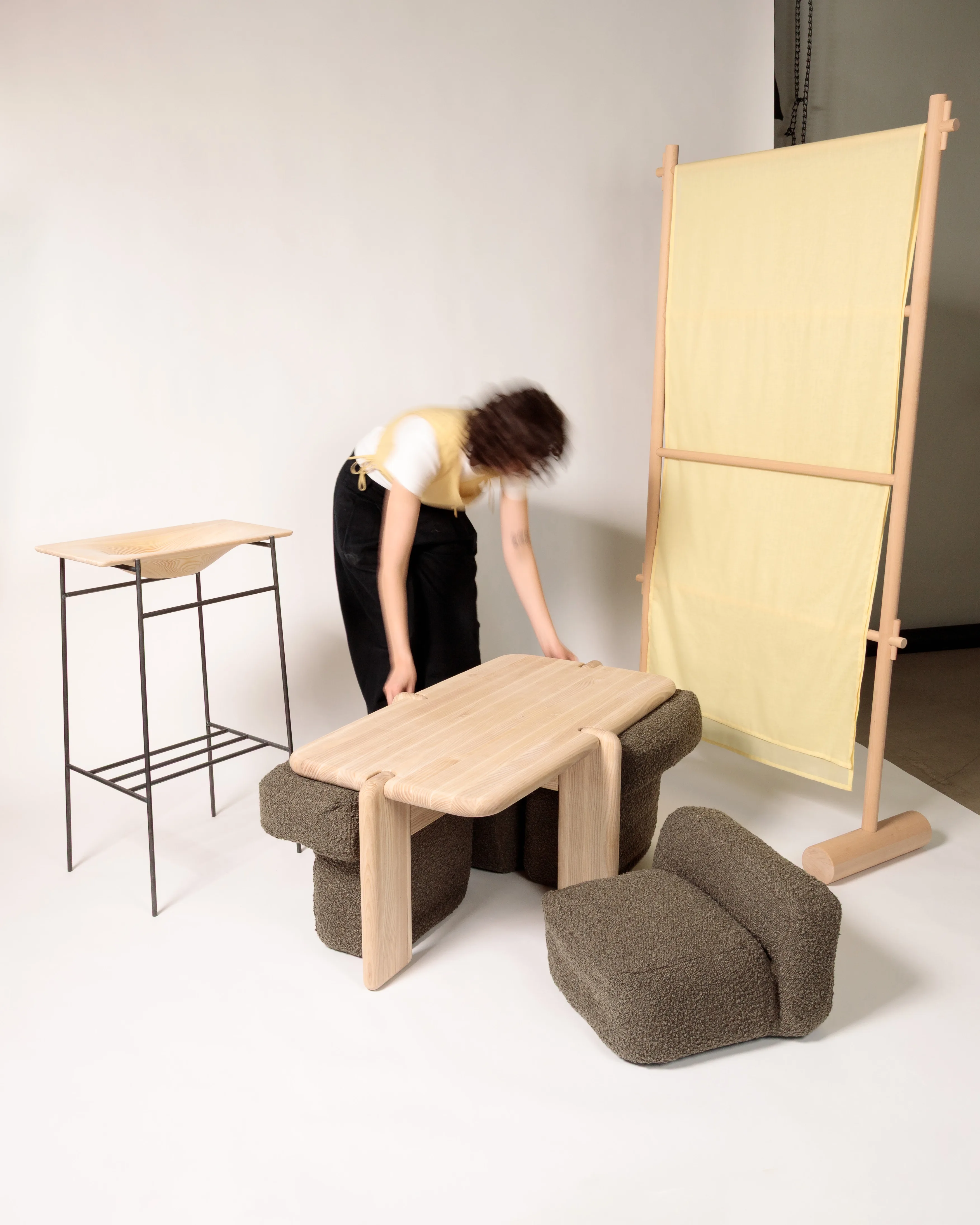 Studio photograph of furniture from Designing for Care—a slender console table, a low table set on bouclé ottomans, and a wood-framed screen with a yellow fabric panel.